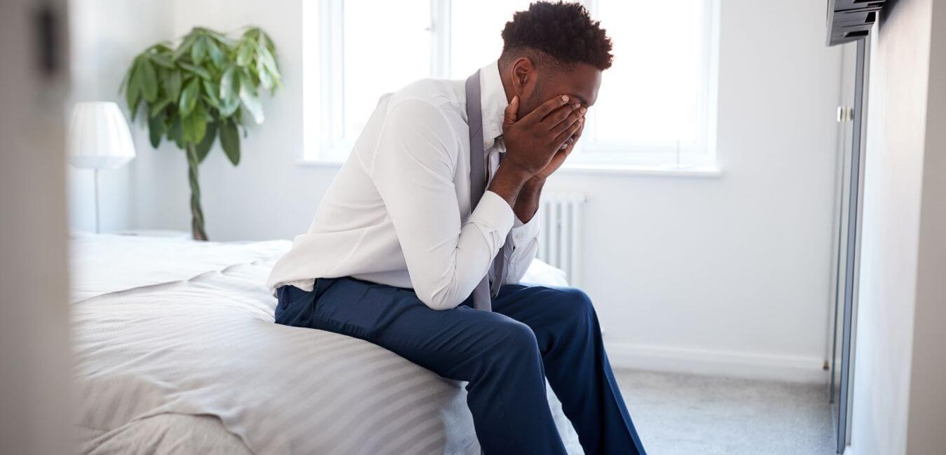 an African American man sits on the edge of a bed with his head in his hands
