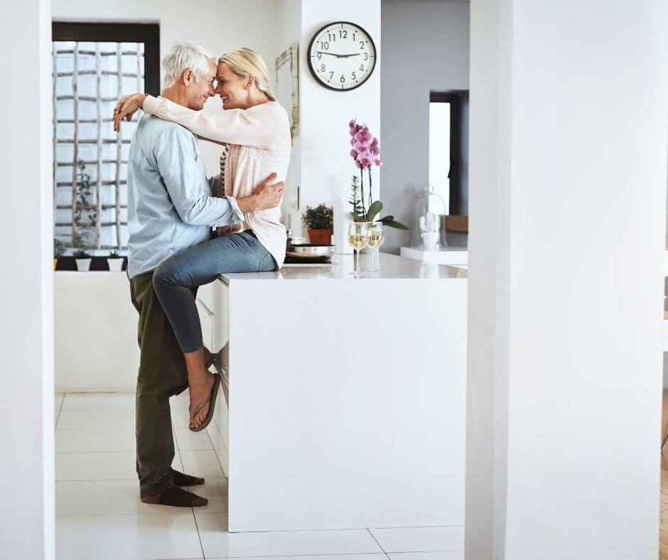 An older woman sits on the kitchen counter while an older man embraces her waist. She has her arms over his shoulders and they're heads are pressed together.