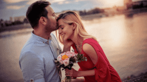 Couple looking sweet and happy while enjoying the sunset together, with the man kissing the woman's forehead as she holds a bouquet of flowers.