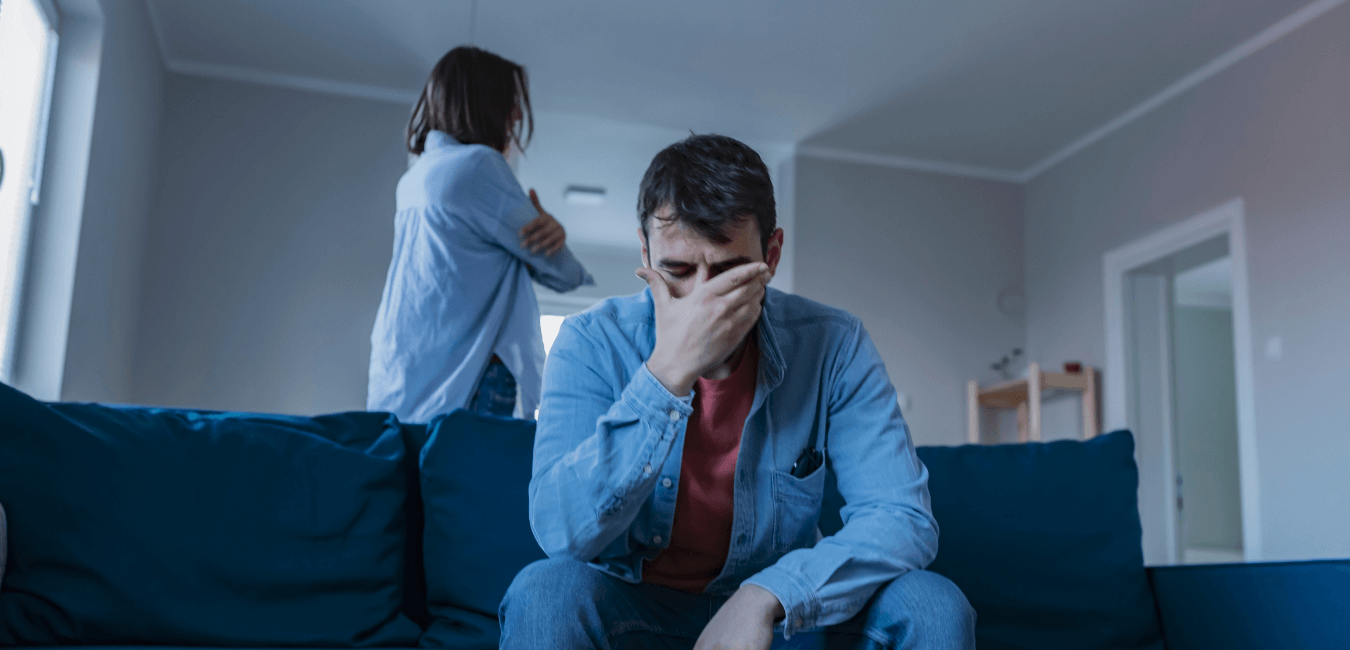A couple arguing, the male sits on the couch head in hands, the woman stands behind the couch arms crossed 