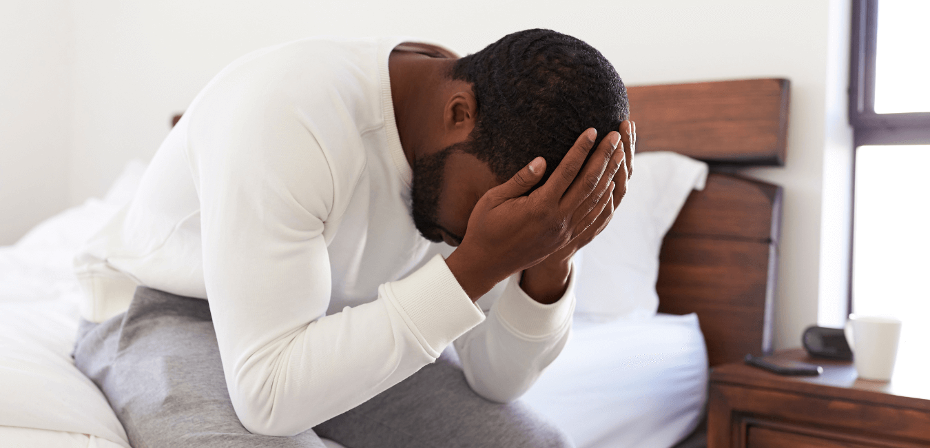 African man sitting on bed with his hand on head looking unhappy