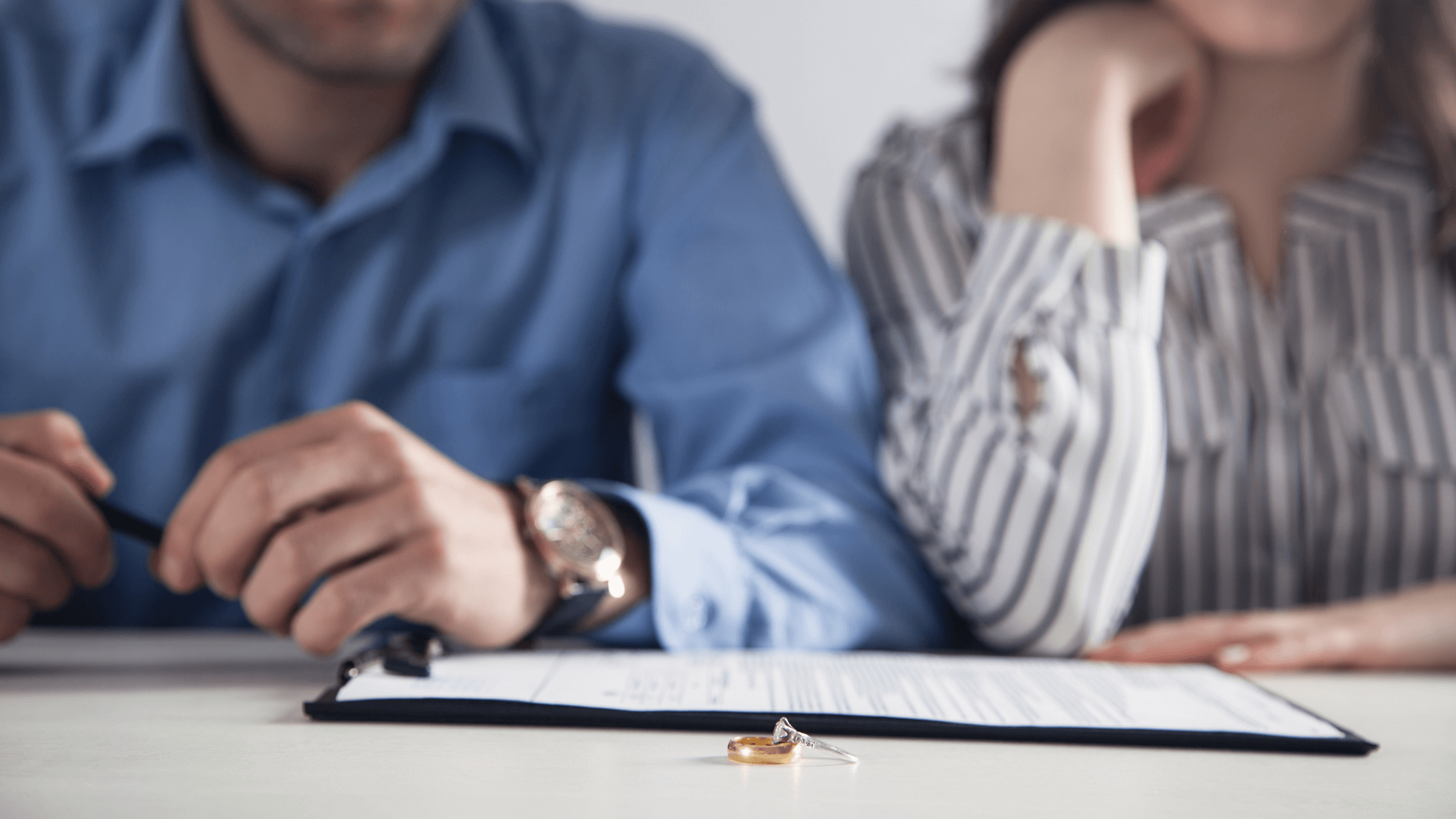 Close-up of wedding ring and divorce papers, couple blurred in background.