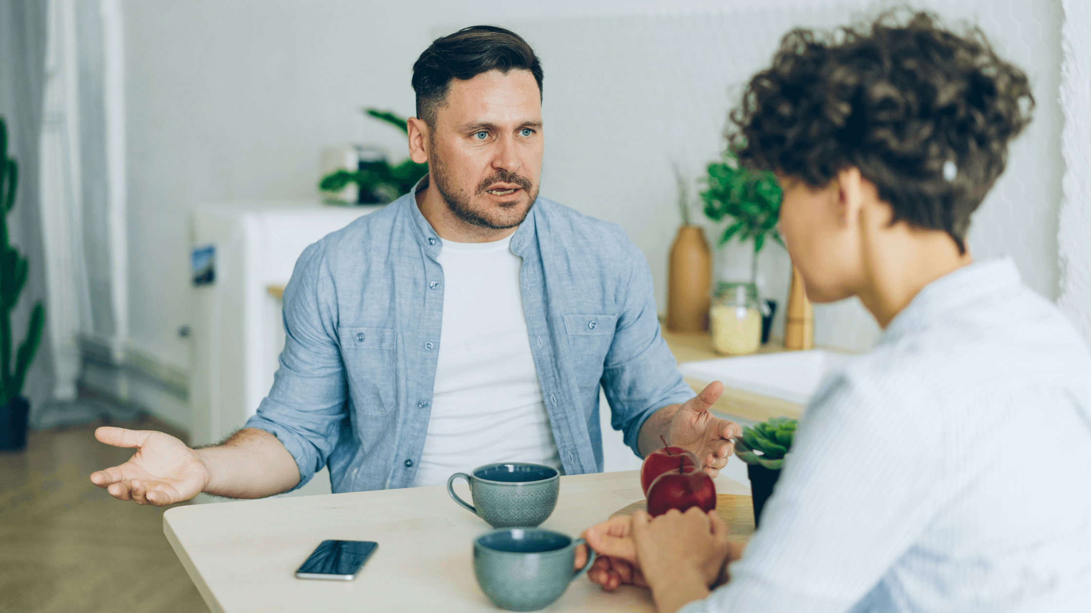 Couple sitting at home table, drinking coffee, man talking with serious expression.