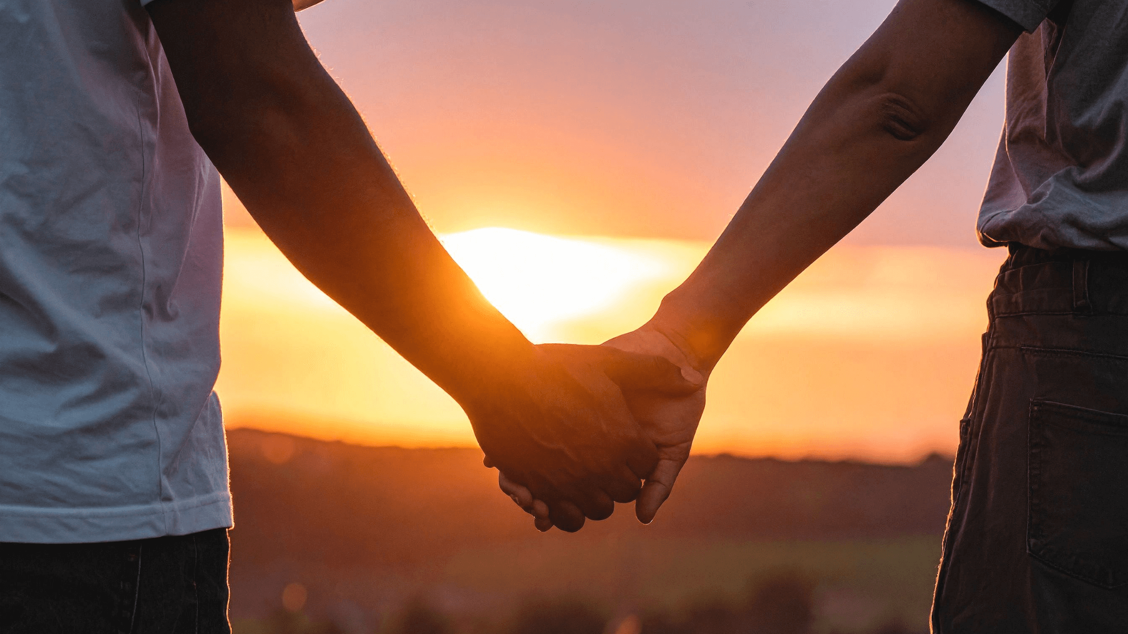 Close-up of a couple holding hands, standing side by side facing the sunset.