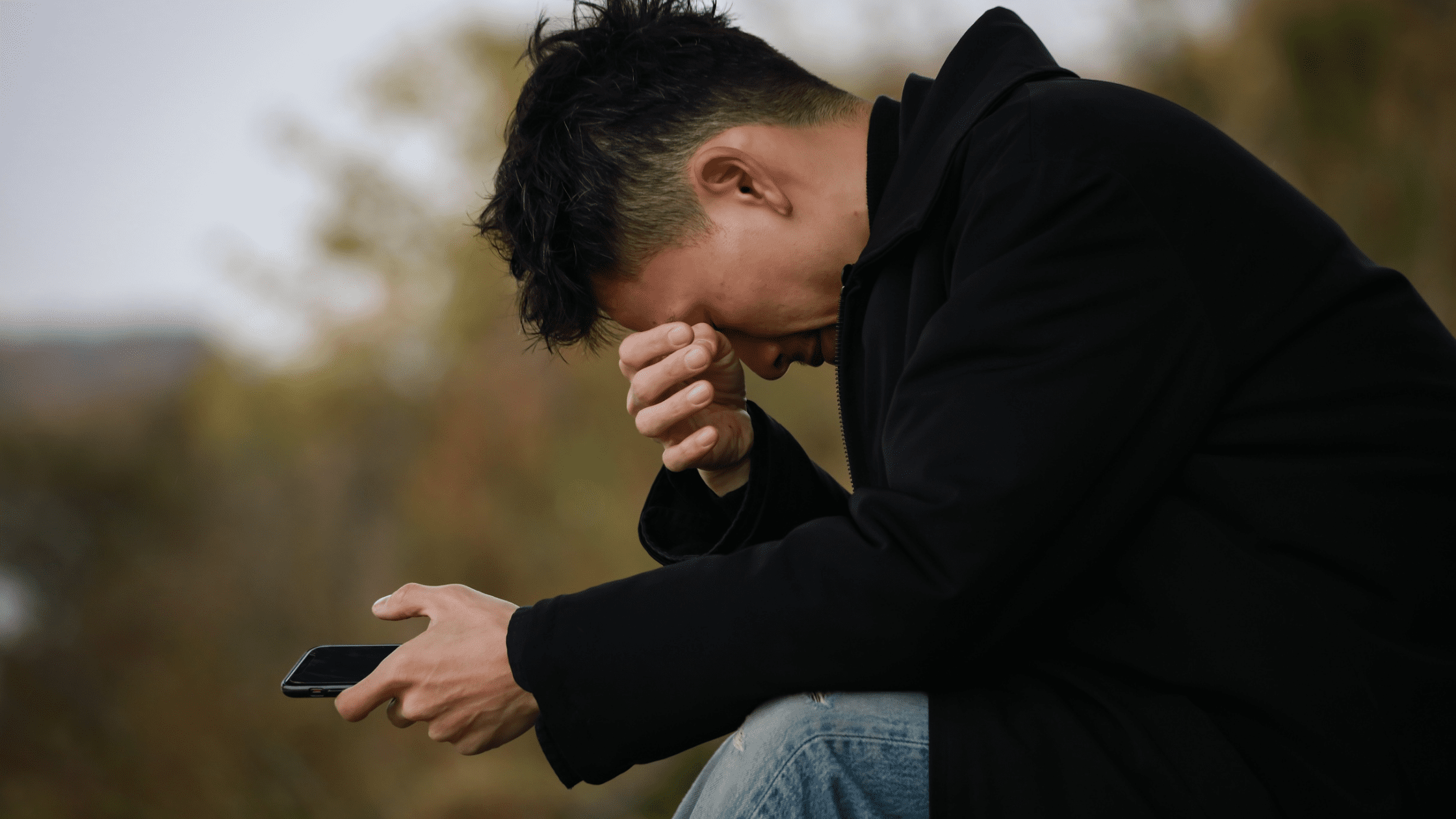 Man sitting outside, holding phone, wiping eyes with hand