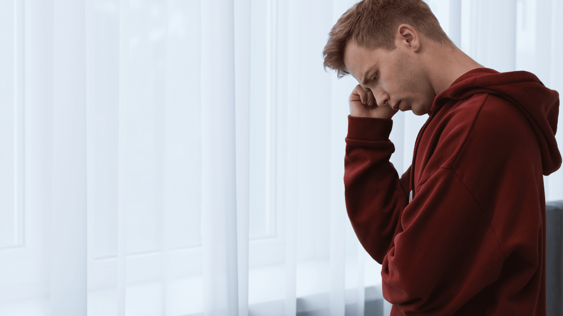 Young man wearing a red sweater standing by the window with a curtain in deep thought.