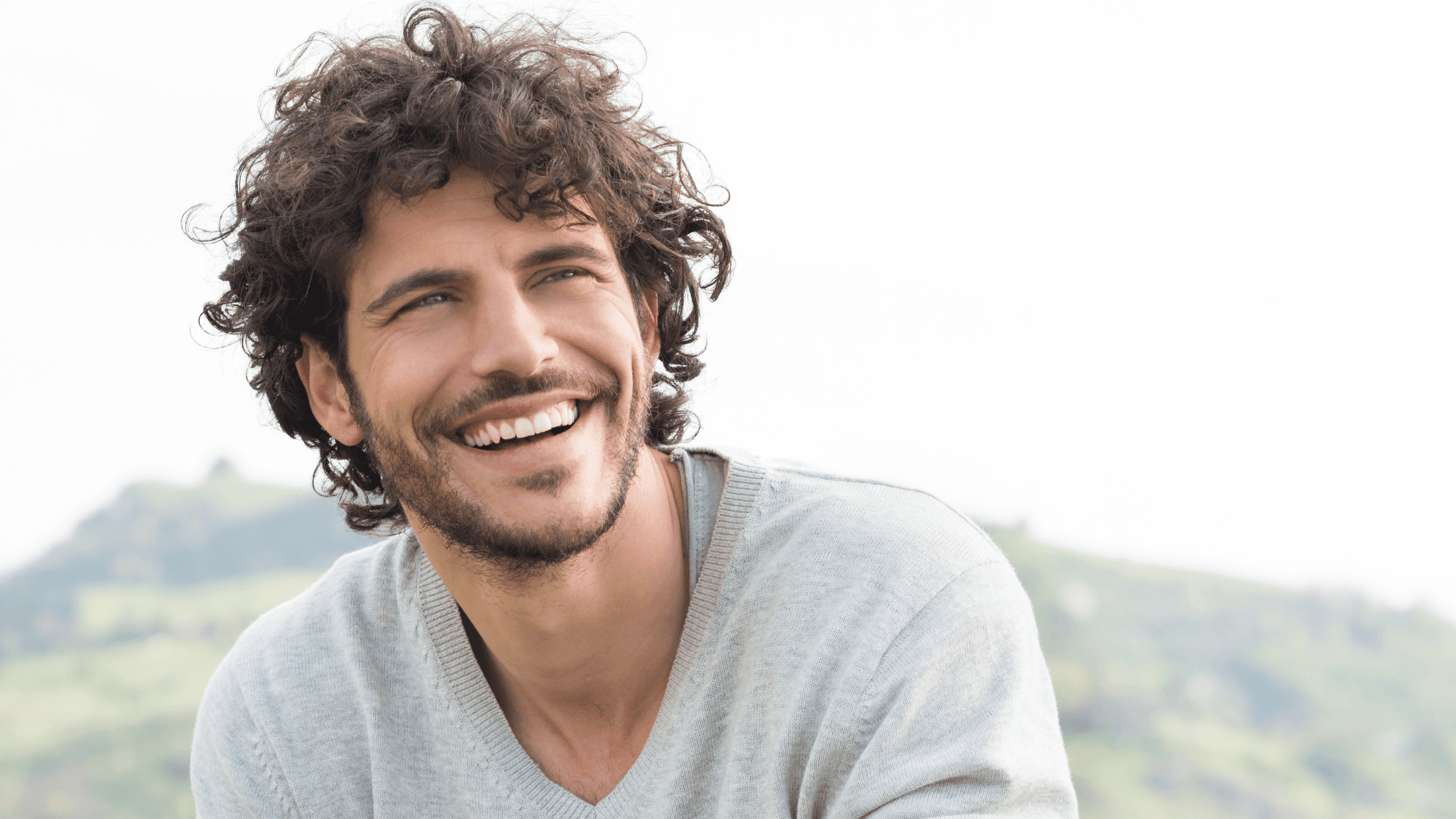 Curly‑haired man smiling outdoors, looking happy.