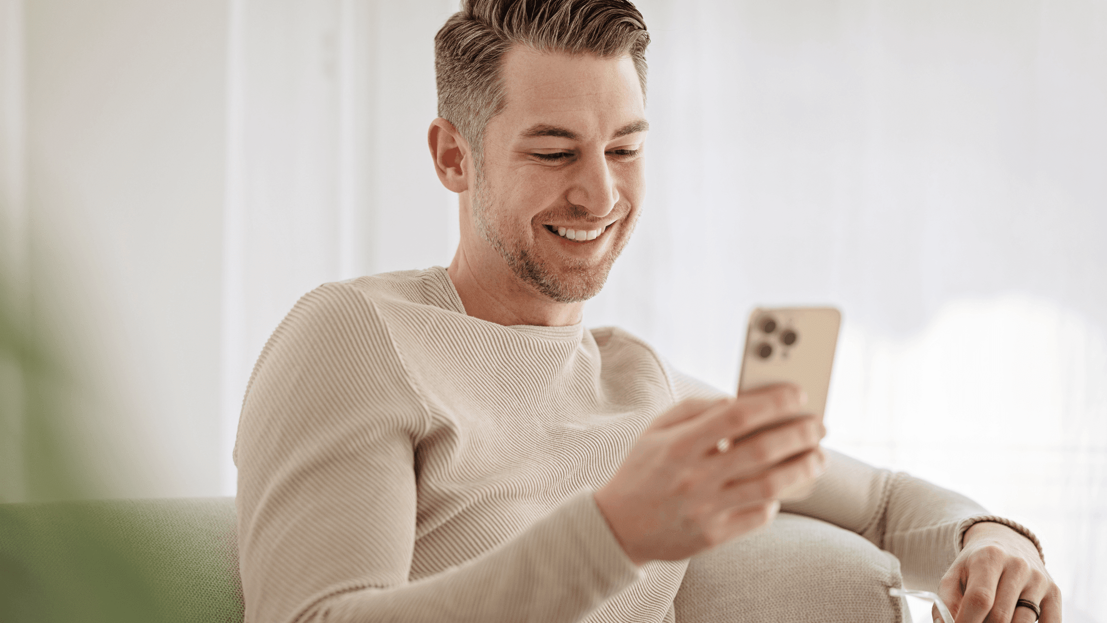 Man sitting comfortably on a couch at home, wearing a sweater, smiling while using a phone.