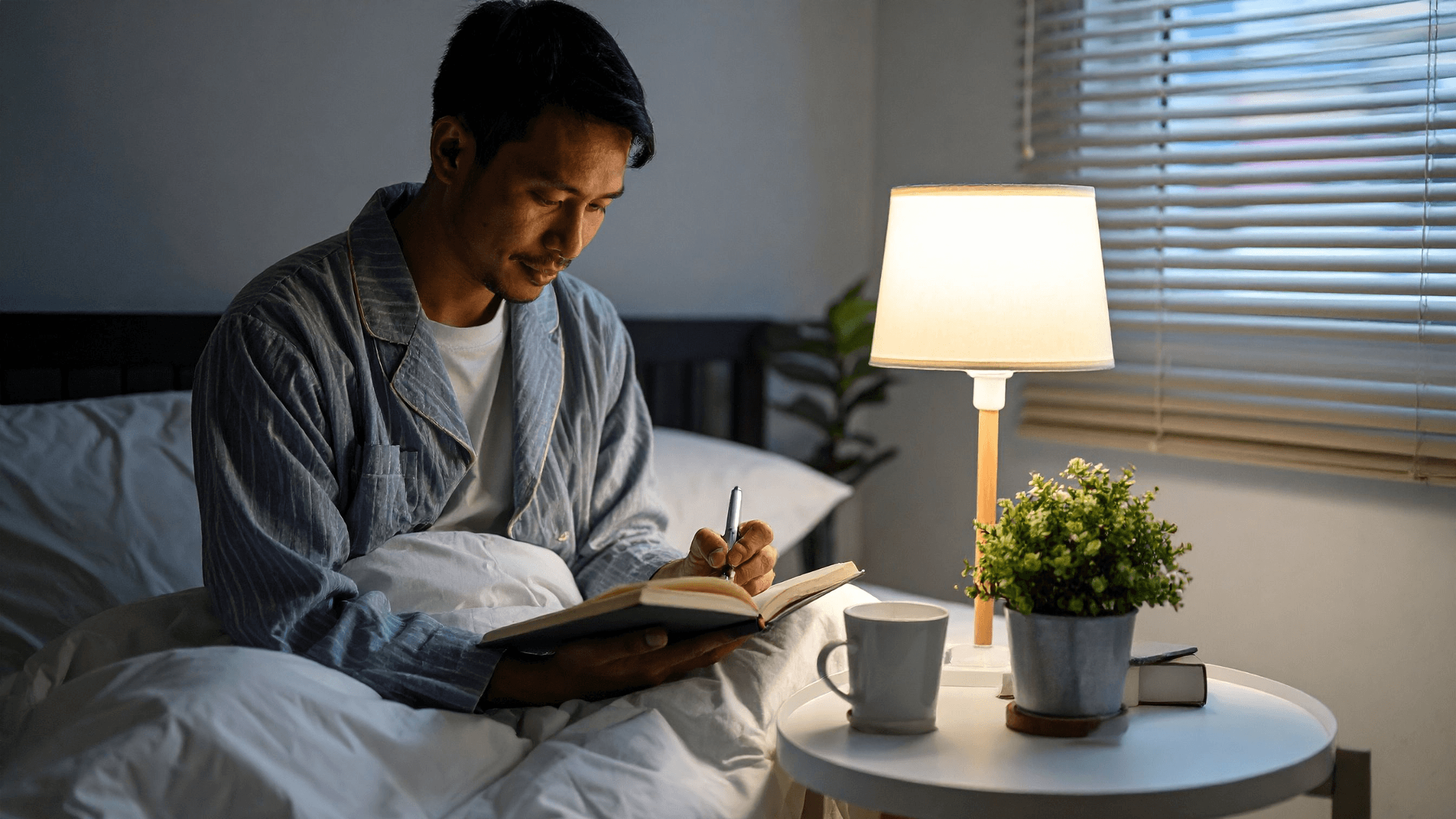 Man in bed wearing sleepwear, writing notes beside shaded lamps.