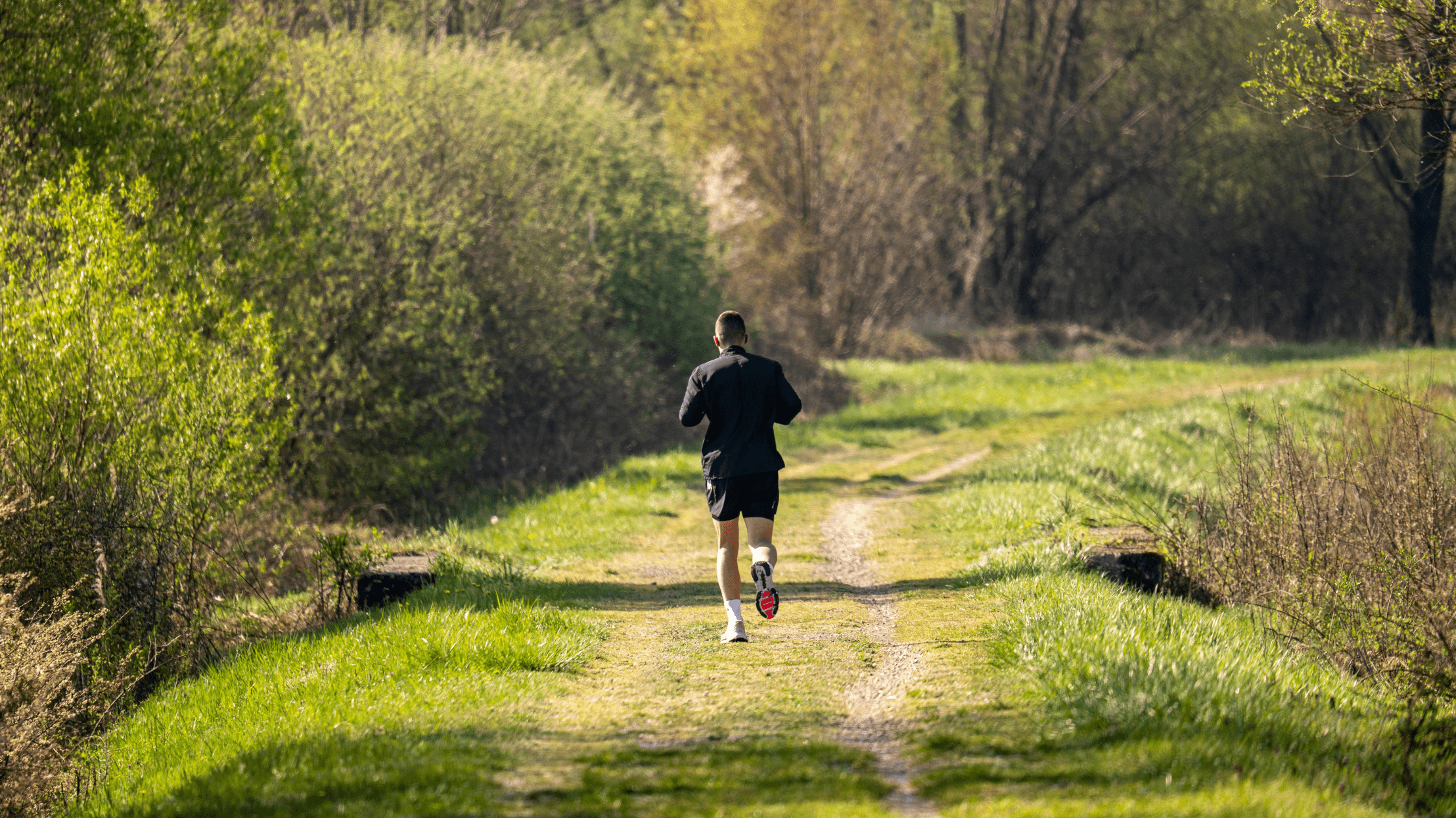 Back shot of a man in black athletic attire running across a lush green outdoor path.