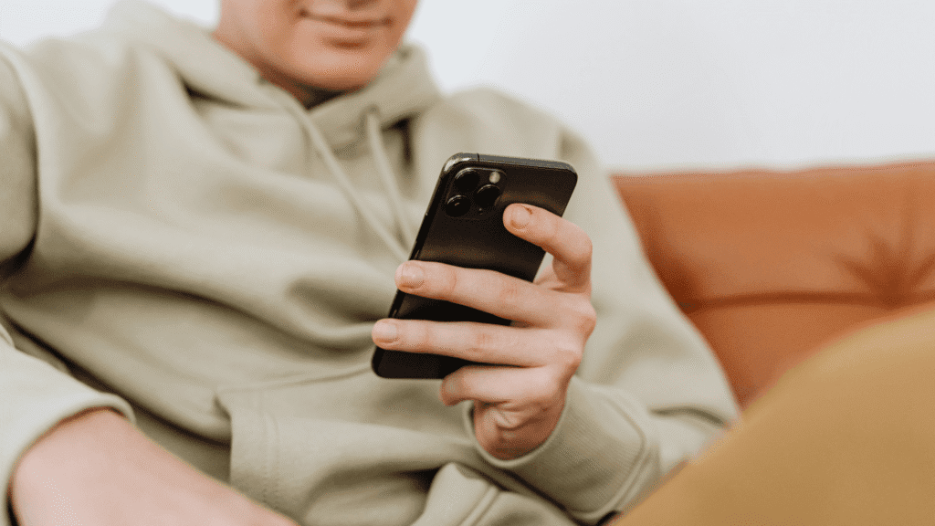 Focused shot of a man of color in a jacket, sitting on a sofa and using a mobile phone, face not visible.