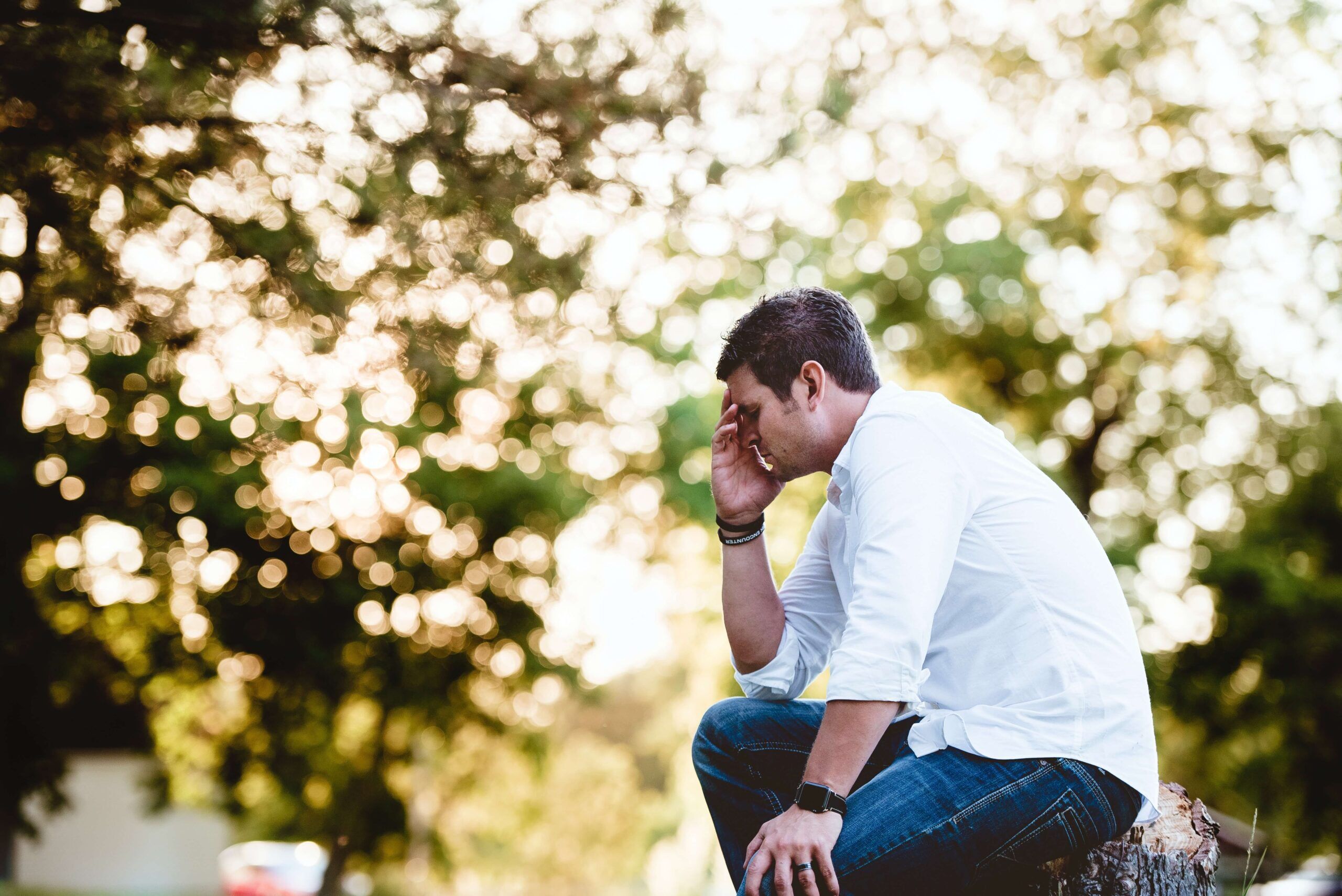 Man sitting outdoors on tree stump looking depressed with his hands on his face