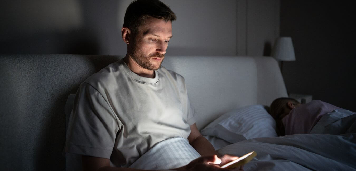 Man using smartphone in a dark bedroom while his wife sleeps beside him.
