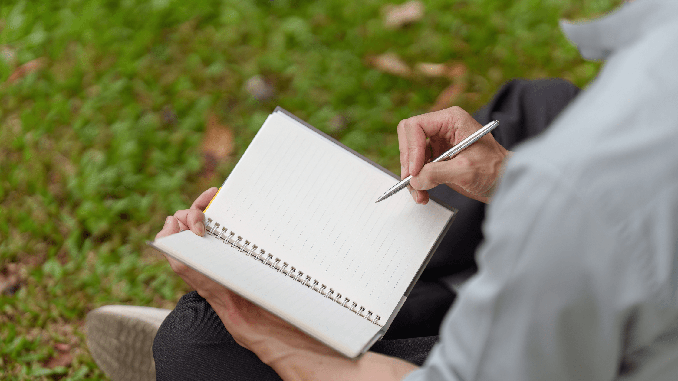 Close-up shot of a man writing in a notebook while sitting in a field.